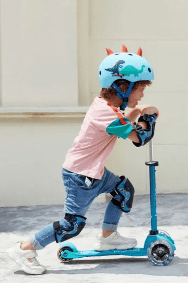 Child riding blue scooter wearing dinosaur helmet and pads