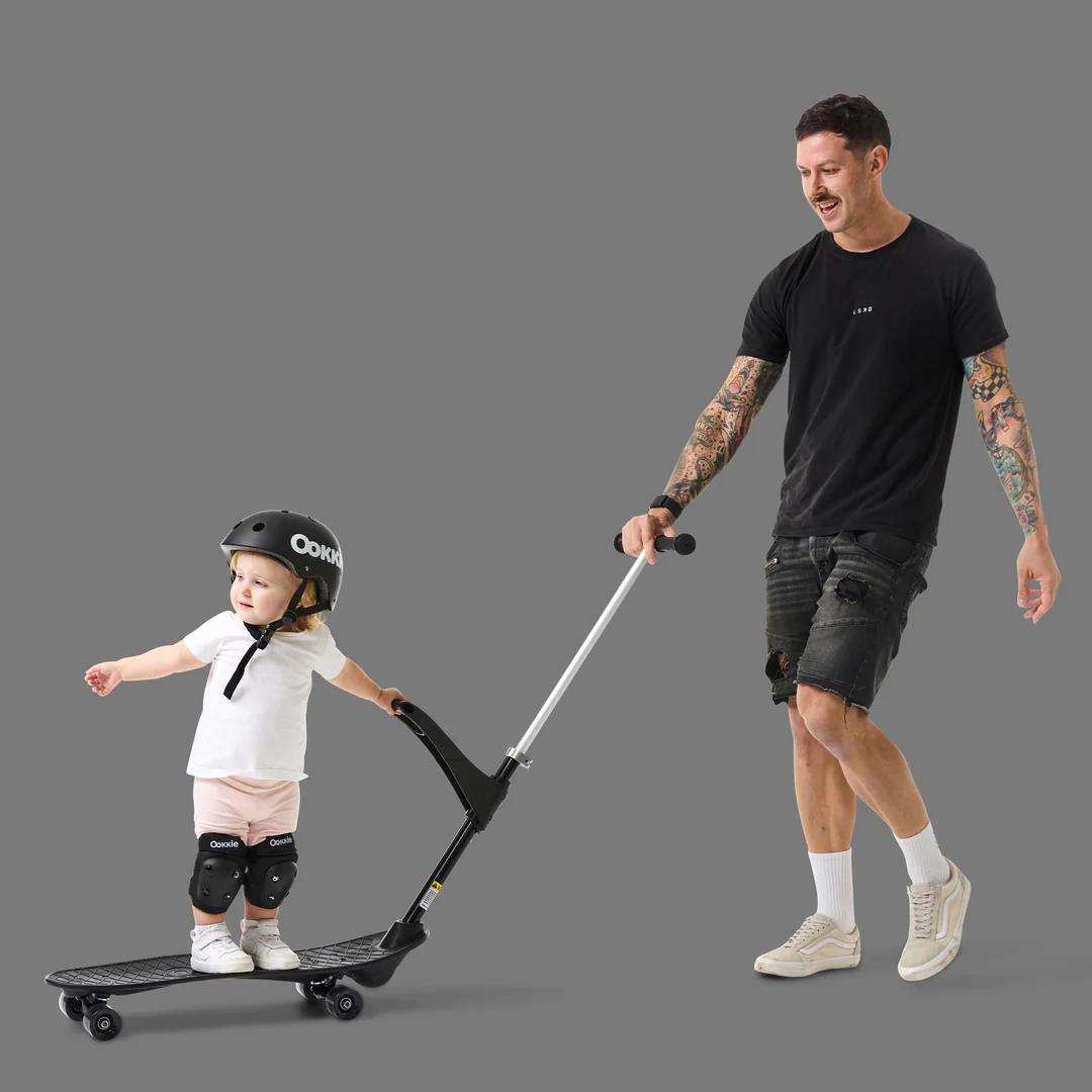 Father guiding toddler on scooter with safety gear on.