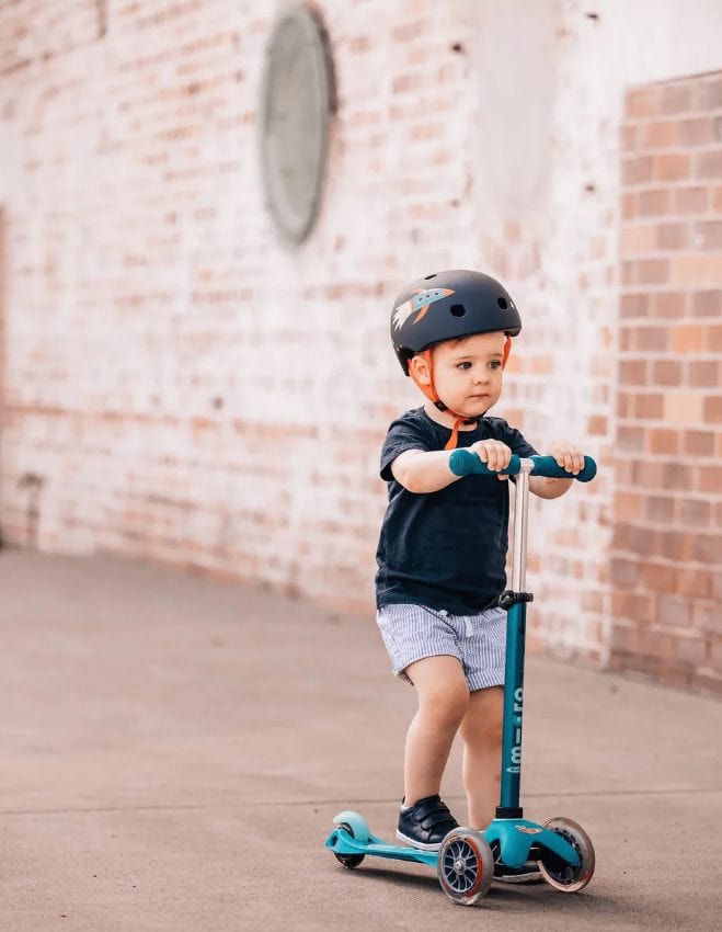 Young child riding teal three-wheel scooter wearing helmet outdoors