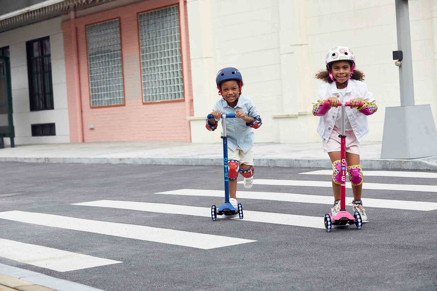 Two smiling children riding scooters across a crosswalk