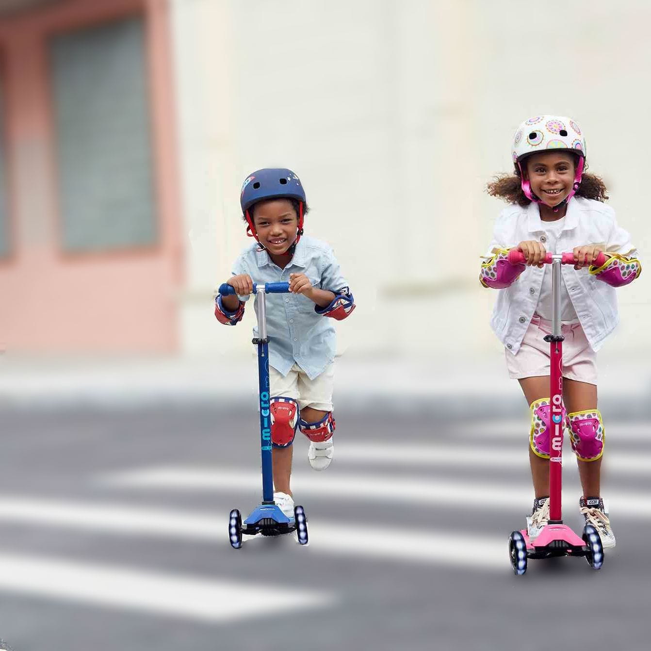 Two smiling children riding scooters across a crosswalk
